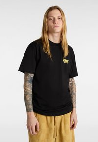 Czarna bawełniana koszulka z krótkim rękawem i okrągłym dekoltem, z żółtym logo "VANS" na piersi. Noszona z jasnymi, żółtymi szortami z sztruksu.