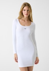 Biała sukienka z długim rękawem o prążkowanej fakturze, z dekoltem w serek oraz małym logo "Guess Jeans" na piersi. Dopasowany krój.
