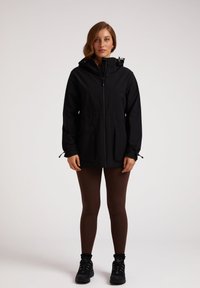 Svart vattentät jacka med huva, framdragkedja, två stora fickor, justerbar dragsko i nederkant, bärs över bruna leggings och svarta skor.