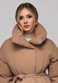 Blondýnka s lehkým make-upem má na sobě béžový zateplený kabát s vysokým límcem a uvázaným páskem, na pozadí jednobarevného šedého pozadí.