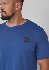 Blauwe katoenen T-shirt met korte mouwen, een ronde hals en een zwart "Discovery" logo met een wereldbolontwerp op de linkerkant van de borst.