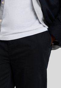 Mörkblå byxor med en slät yta, kombinerade med en vit skjorta och en svart jacka. Jackan har ett randigt foder och knäppning fram.