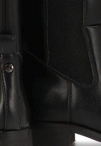 Bota de cuero negro con panel elástico, textura suave, suela resistente y detalle metálico con el logotipo cerca del tobillo.