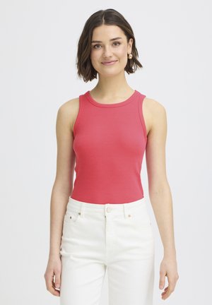 Femme aux cheveux courts bruns portant des boucles d'oreilles dorées en forme de créoles, un haut rouge sans manches et un pantalon blanc taille haute, debout et souriante.