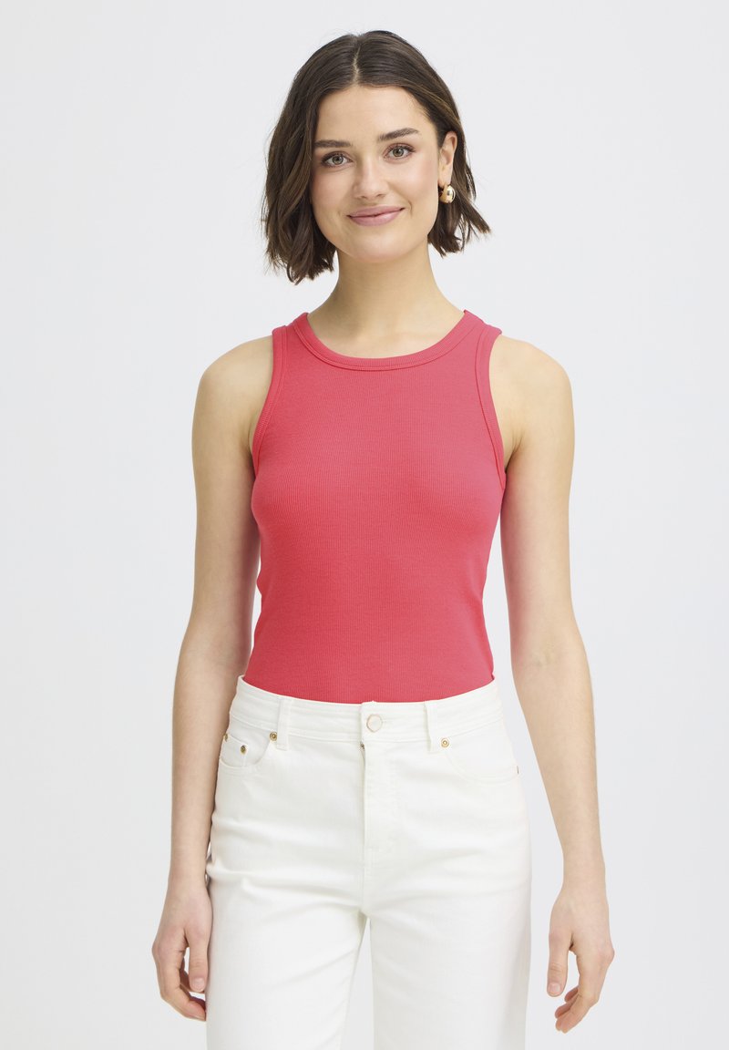 Kobieta o krótkich brązowych włosach, nosząca złote kolczyki w kształcie koła, czerwoną bezrękawnikową bluzkę i białe spodnie z wysokim stanem, stojąca i uśmiechnięta.