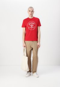 T-shirt en coton rouge avec le logo graphique blanc "JOOP! JEANS" centré sur la poitrine, associé à un pantalon beige et un sac fourre-tout crème. Baskets blanches.