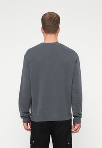 Uomo con capelli corti castani che indossa un maglione di maglia grigia e pantaloni neri, rivolto di spalle contro uno sfondo chiaro e semplice.