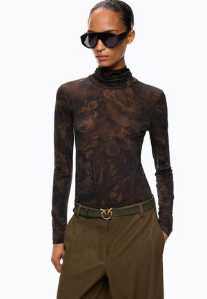 Mujer con un jersey de cuello vuelto de manga larga con estampado floral oscuro, pantalones verde oliva con un cinturón a juego con hebilla dorada y gafas de sol negras de gran tamaño.