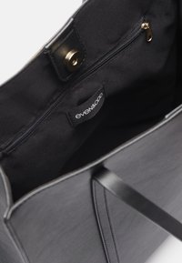 Interiér černé tašky na rameno obsahuje zipovou kapsu, hladkou tkaninu a značkovou visačku. Taška má ploché, pevné popruhy a čisté švy.