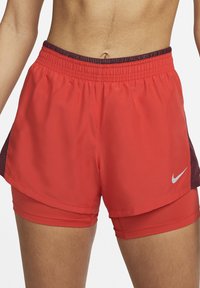 Pantalones cortos deportivos rojos con mallas de compresión incorporadas y un pequeño logo blanco de Nike en el muslo derecho, llevados por una persona.