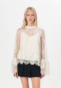 Blusa in pizzo trasparente con maniche lunghe e svasate. Presenta motivi floreali, un collo alto e un bordo in pizzo scallopato all'orlo.
