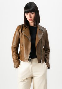 Donna che indossa una giacca di pelle marrone, una maglietta nera e pantaloni crema, in piedi con le mani nelle tasche su uno sfondo bianco semplice.