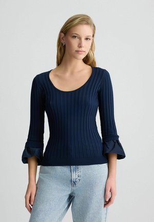 Marineblaues ribbelstrick Oberteil mit einem runden Ausschnitt und 3/4-Ärmeln mit ausgestellten Bündchen, kombiniert mit hellblauen Jeans.