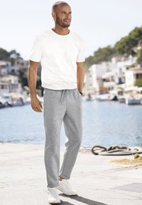 Weißes Baumwoll-T-Shirt und graue Sweatpants mit Kordelzug. Das Model trägt weiße Sneaker. Der Hintergrund zeigt eine Uferlandschaft und Gebäude.