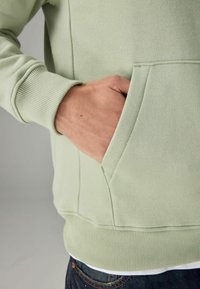 Sudadera verde claro con un bolsillo canguro, puños y dobladillo acanalados, que presenta una textura suave y un diseño sin costuras.