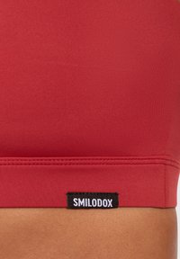 Top corto atlético rojo, hecho de una tela suave, con una banda ancha y una etiqueta negra con "SMILODOX" impresa en blanco.
