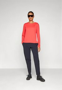 Crvena majica dugih rukava u kombinaciji s tamno plavim hlačama, s opuštenim krojem. Dodaci uključuju sunčane naočale i crne cipele. Glatka tekstura tkanine.