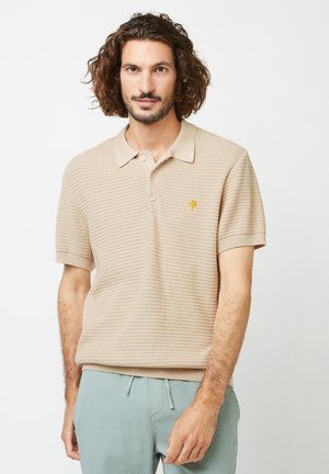Beige gebreide polo-shirt met een kraag, korte mouwen en een klein gouden logo op de borst. Gestructureerde stof, ribbelboorden.