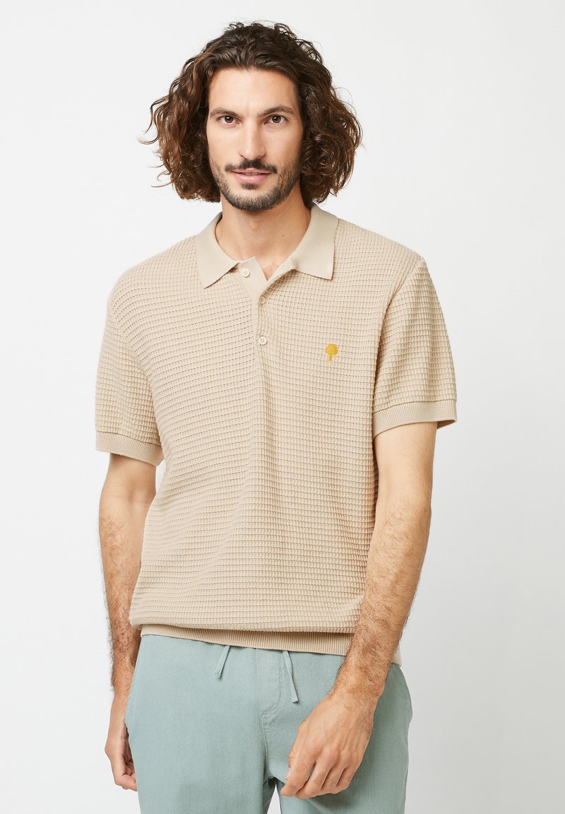 Polo en tricot beige avec un col, des manches courtes et un petit logo doré sur la poitrine. Tissu texturé, poignets côtelés.