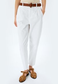 Pantaloni di cotone bianchi con pieghe e tasche laterali, abbinati a scarpe in suede marrone con un design intrecciato e cinturino alla caviglia.
