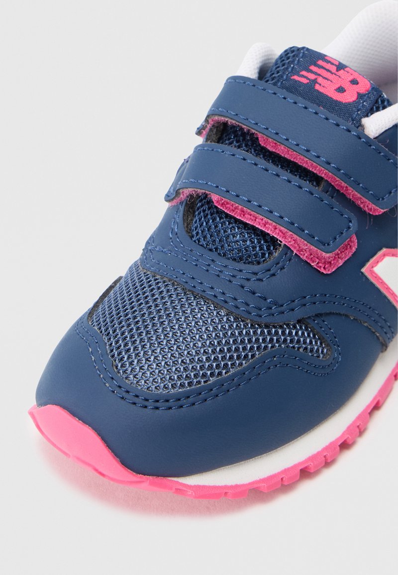 Zapatilla deportiva azul con parte superior de malla y cuero suave, doble cierre de tiras de velcro, acentos rosas y una suela de goma texturizada en rosa.