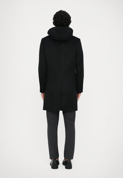 Abrigo negro con capucha, hecho de una mezcla de lana. Presenta un corte recto, un diseño de longitud media y mangas entalladas. Combinado con pantalones grises.