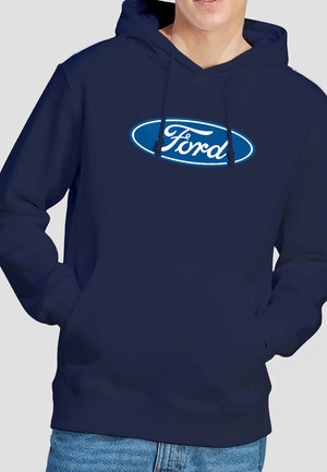 Person wearing navy blue hoodie with white and blue oval Ford logo on chest, hands in front pouch pocket, paired with blue jeans.