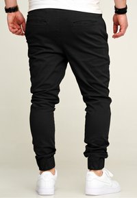 Black joggers made from a stretchy fabric feature two back pockets, elasticated cuffs, and are paired with white sneakers.