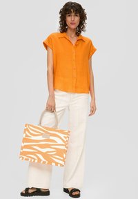 Camicia arancione in lino a maniche corte con bottoni, abbinata a pantaloni larghi color crema e una borsa a tracolla con manici beige e stampa zebrata arancione.