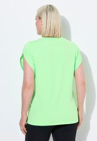 Grøn kortærmet skjorte lavet af et glat, strækbart stof, med en afslappet pasform og rund kant, båret med sorte leggings.