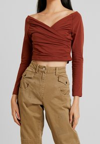 Rostroter, langärmeliger Croptop mit Wickeldesign und gerafftem Stoff am Dekolleté, kombiniert mit khakifarbenen Hosen mit Patchdetails und Taschen.