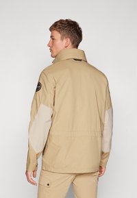 Giacca beige con colletto alto, pannelli delle spalle in beige chiaro contrastante e una toppa sul braccio sinistro. Presenta una vestibilità comoda e cuciture visibili.