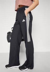 Svarta träningsbyxor med vita vertikala ränder och Adidas-logotyp, med en avslappnad passform och sidofickor. Bärs med svarta skor.