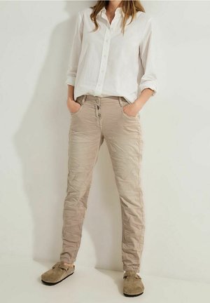 Femme portant un pantalon beige ajusté avec des boutons et des poches, une chemise blanche à boutons avec les manches retroussées, et des chaussures beiges sans lacets.