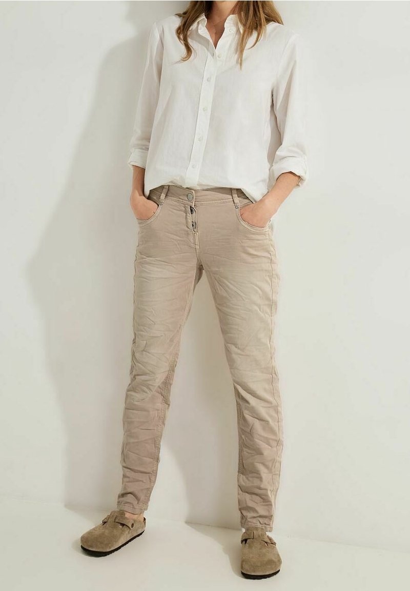 Femme portant un pantalon beige ajusté avec des boutons et des poches, une chemise blanche à boutons avec les manches retroussées, et des chaussures beiges sans lacets.