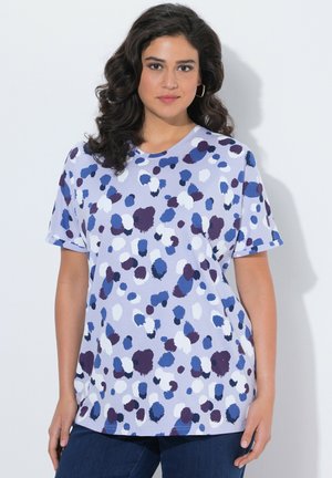 Kobieta o ciemnych, kręconych włosach, ubrana w krótkim rękawem, luźną, lawendową koszulę z niebieskimi, białymi i ciemnofioletowymi abstrakcyjnymi plamami, w zestawieniu z ciemnymi dżinsami.