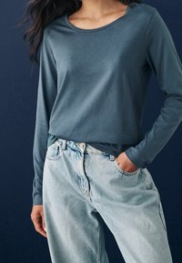 Donkerturquoise top met lange mouwen, gemaakt van zachte stof, in combinatie met lichtblauwe, high-waisted denim jeans. Heeft een ronde halslijn en een relaxte pasvorm.