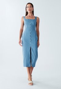 Zila kleita ar pogām priekšpusē, izgatavota no gludas auduma, ar kvadrātveida kakla izgriezumu, plašiem lencēm un priekšējo šķēlumu kā papildus detaļu.