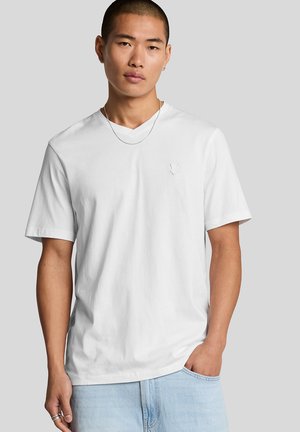 Homme portant un t-shirt blanc uni à col en V, un jean bleu clair, un collier fin en argent et des bagues en argent, debout avec une main dans la poche.