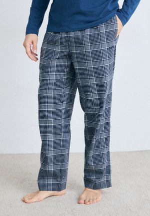 Person steht barfuß auf dem Teppich und trägt karierte navy und weiße Hosen sowie ein langärmliges navy blaues Hemd, mit einer Hand in der Tasche.