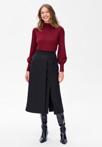 Uśmiechnięta kobieta z luźnymi falującymi włosami, ubrana w bordową bluzkę z długim rękawem, czarną midi spódnicę z rozcięciem z przodu oraz błyszczące czarne botki za kolano.