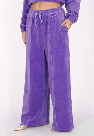 Frau trägt lockere violette Velourhose mit genieteten Taschen und einem Kordelzug in der Taille, kombiniert mit goldenen spitzen Schuhen.