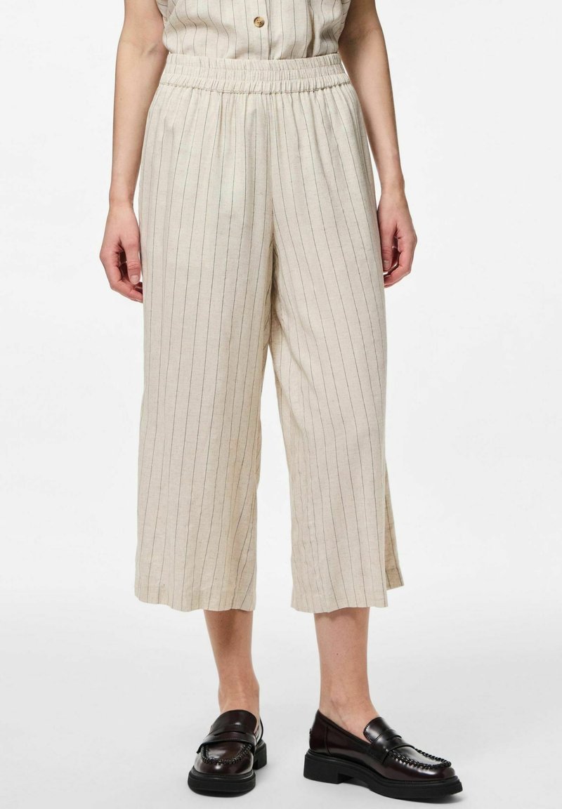 Pantalons larges coupés beiges à fines rayures verticales portés avec des mocassins noirs épais par une personne debout.