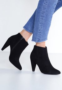 Mustat mokkanahkaiset nilkkurit, joissa on kapeneva korkea korko, sivuvetoketju ja sileä pinta, yhdistettyinä lyhyisiin sinisiin farkkuihin.