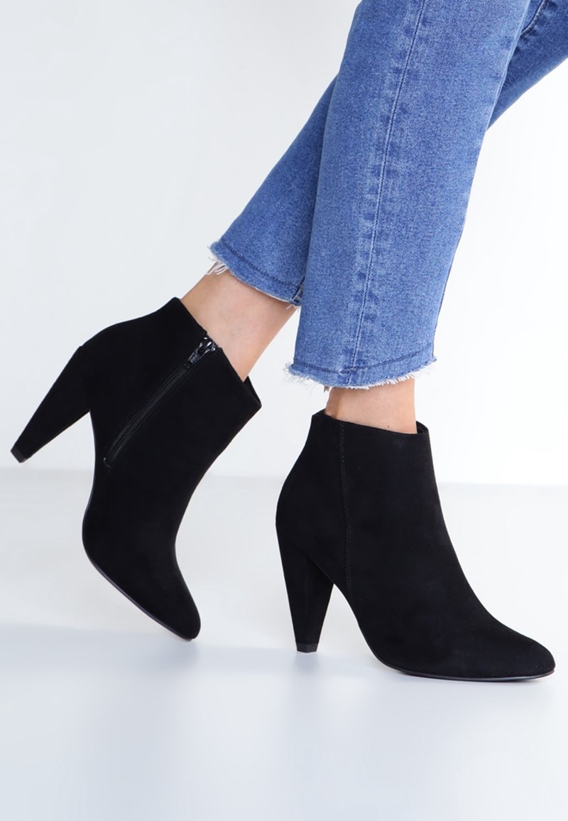 Mustat mokkanahkaiset nilkkurit, joissa on kapeneva korkea korko, sivuvetoketju ja sileä pinta, yhdistettyinä lyhyisiin sinisiin farkkuihin.