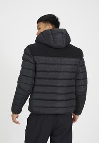 Chaqueta acolchada negra con capucha, que presenta paneles acolchados y un yugo de hombro negro texturizado. Tejido suave y diseño ajustado.