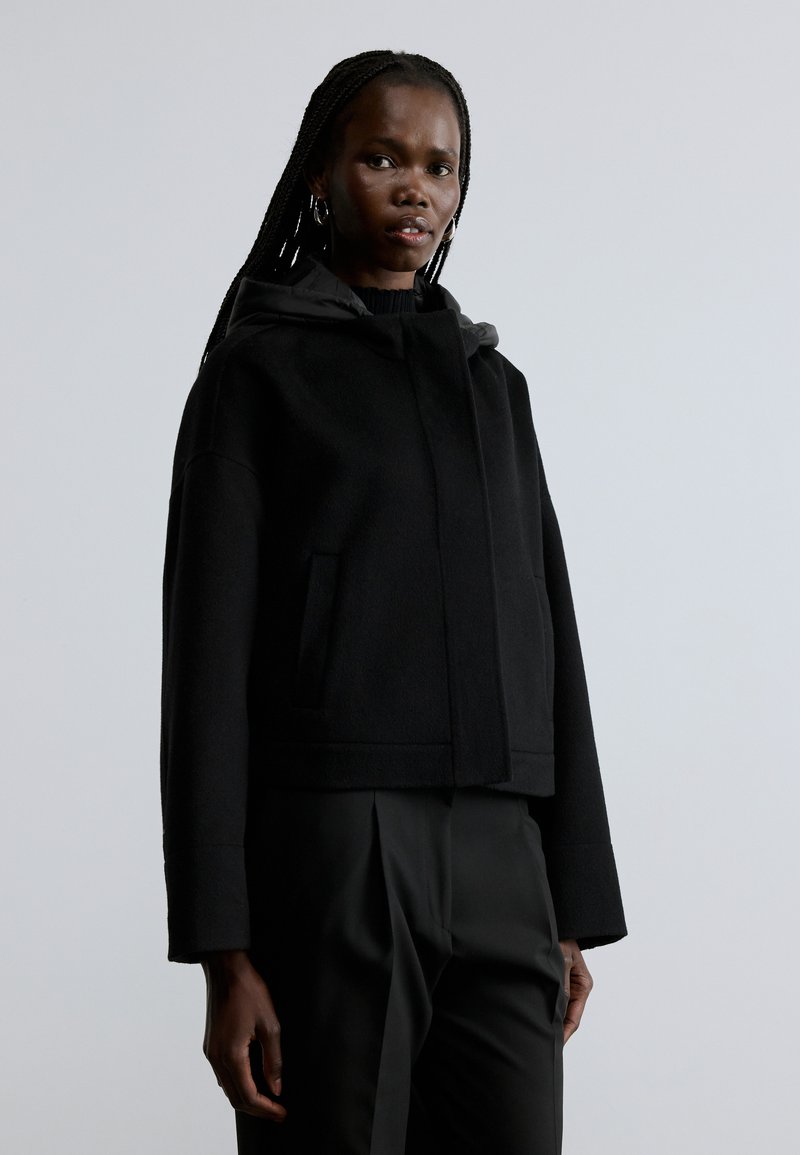 Veste noire courte avec capuche, dotée d'une texture douce, de poches avant et d'un design minimaliste. Associée à un pantalon noir.