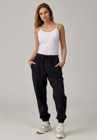 Zwarte sweatpants met elastische boorden, trekkoord taille; witte tanktop met dunne banden. Model draagt lichtgekleurde sneakers.