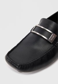 Μαύρες δερμάτινες μοκασίνες με επίπεδη σιλουέτα, διαθέτοντας μεταλλική ασημί λεπτομέρεια και καλοραμμένες ραφές κατά μήκος των άκρων.