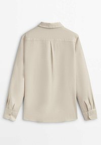 Chemise beige à manches longues avec poignets boutonnés et pli central au dos, présentée sur un fond blanc.